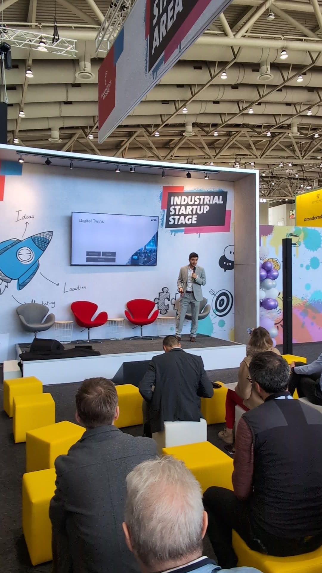 Vortrag über digitale Zwillinge auf der Industrial Startup Stage, das Publikum sitzt auf gelben Sitzwürfeln